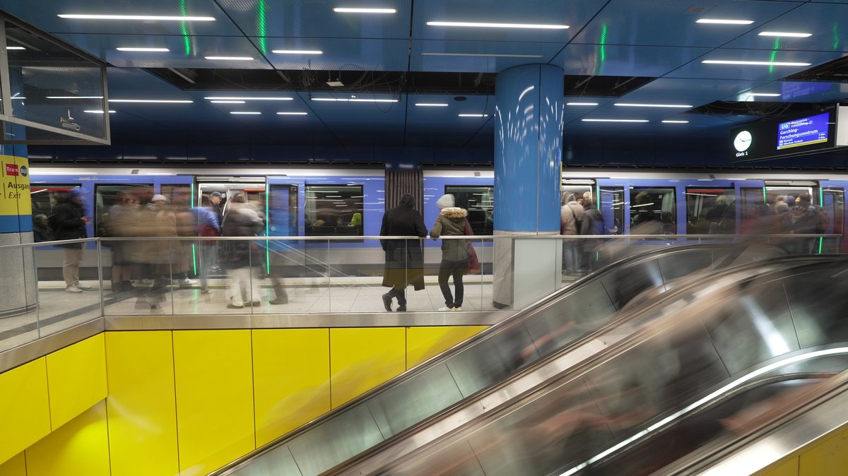 Im U-Bahnhof steht eine U-Bahn, davor sieht man verwischt Menschen warten und auf einer Rolltreppe fahren
