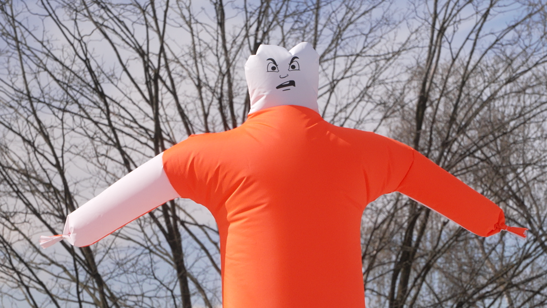 Vor den Bäumen sieht man einen aufgeblasenen, etwas unförmigen Oberkörper in den Signalfarben rot und weiß. Der aufgemalte Gesichtsausdruck ist grimmig.