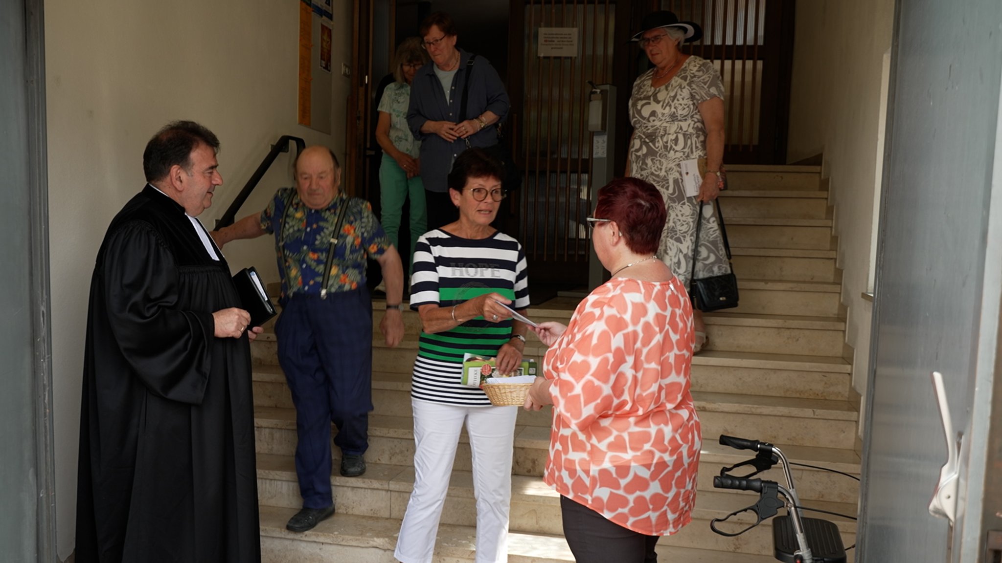 Kirchenbesucher erhalten nach dem Gottesdienst Umschläge an der Eingangstür; der Pfarrer steht daneben in seinem schwarzen Talar und beobachtet die Szene.