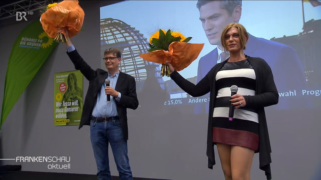 Sascha Müller (links) und Tessa Ganserer jubeln mit Blumen in der Hand auf einer Bühne.