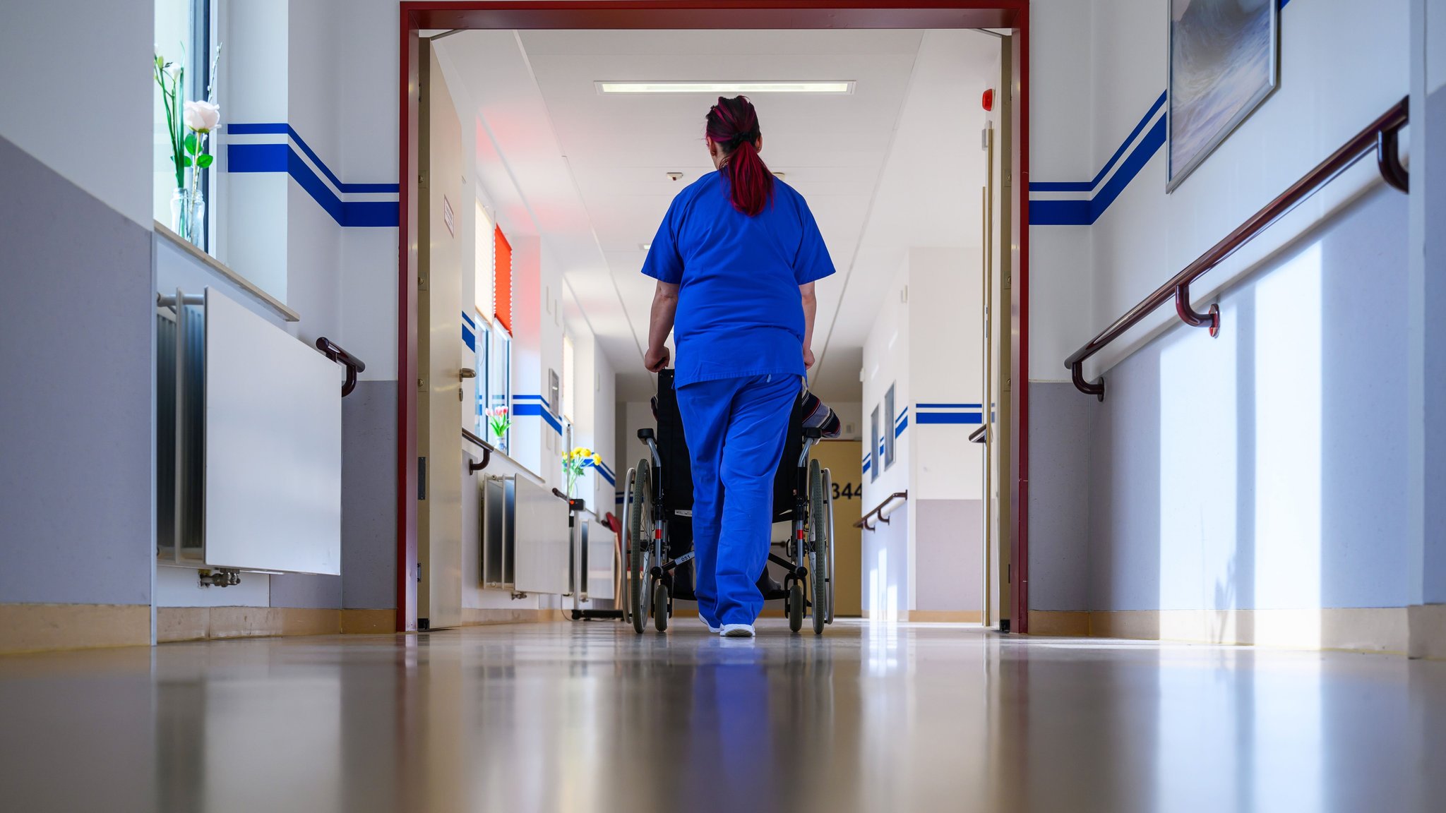Eine Pflegerin in einem Krankenhaus schiebt einen Patienten im Rollstuhl über die Station. (Symbolbild) | Bild: dpa-Bildfunk/Hendrik Schmidt Eine Pflegerin in einem Krankenhaus schiebt einen Patienten im Rollstuhl über die Station. (Symbolbild)