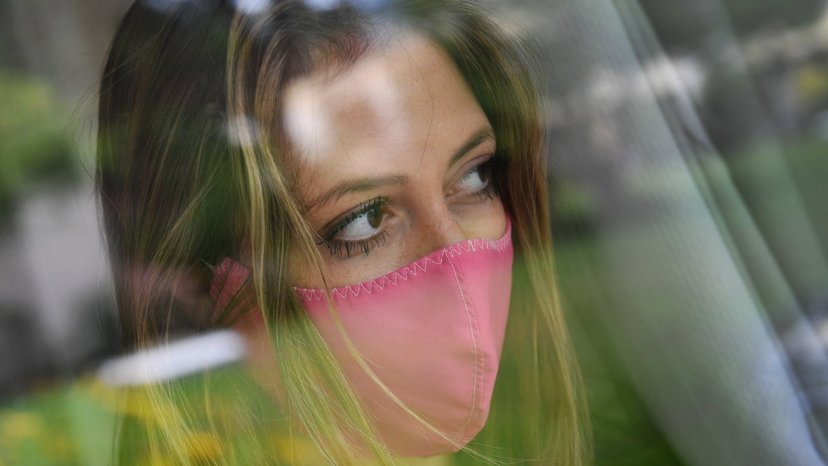Eine junge Frau mit pinker Schutzmaske steht am Fenster in einem Zimmer und blickt wehmuetig, traurig in die Ferne.