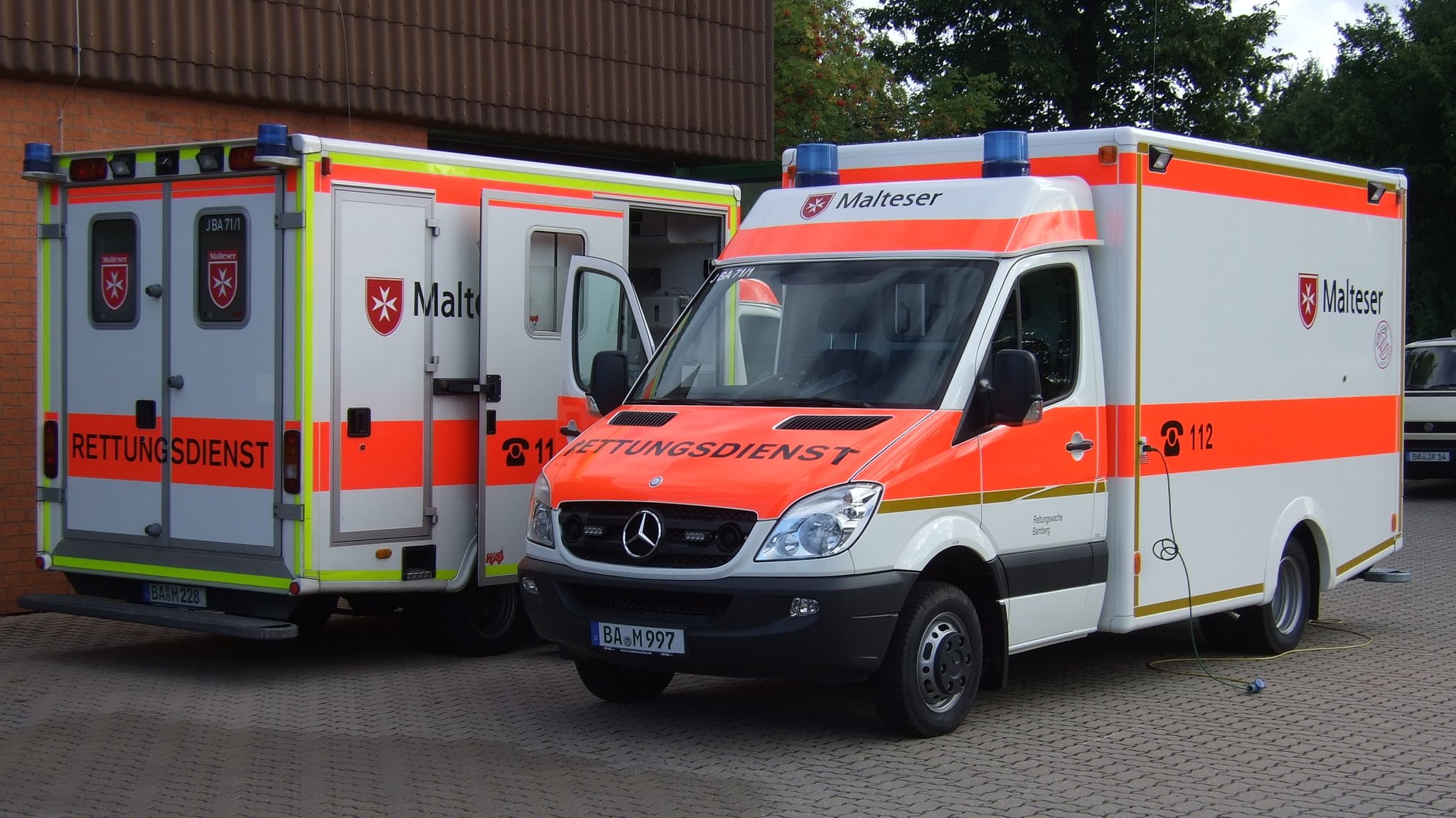 Zwei Rettungstransporter der Malteser parken auf einem Hof. Auf dem Wagen steht in schwarzer Schrift auf rotem Hintergrund "Rettungsdienst". Auf der Seite des Krankenwagens steht auf rotem Hintergrund 112. Außerdem steht auf beiden Transportern Malteser, daneben ist das Zeichen der Hilfsorganisation abgebildet.