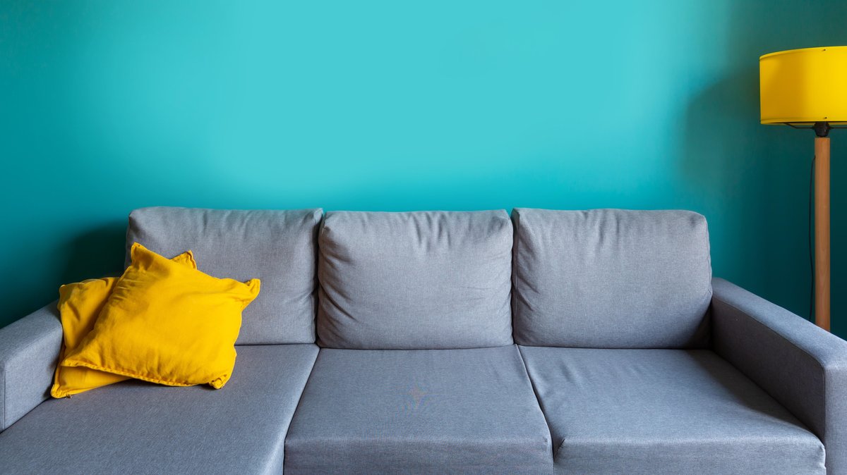 Ein graues Sofa mit gelben Kissen und gelber Lampe vor einer türkisfarbenen Wand (Symbolbild)