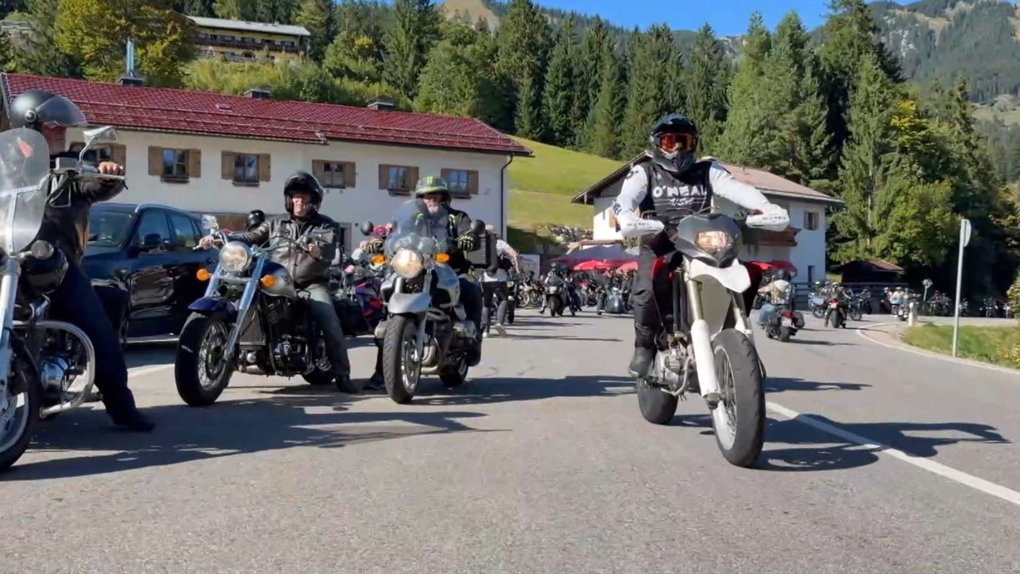 Mehrere Männer auf Motorrädern fahren auf einer Straße. Im Hintergrund sind zwei Gebäude zu sehen. Dahinter befinden sich Hügel und Wälder. | Bild: BR/Boris Berg Mehrere Männer auf Motorrädern fahren auf einer Straße. Im Hintergrund sind zwei Gebäude zu sehen. Dahinter befinden sich Hügel und Wälder.