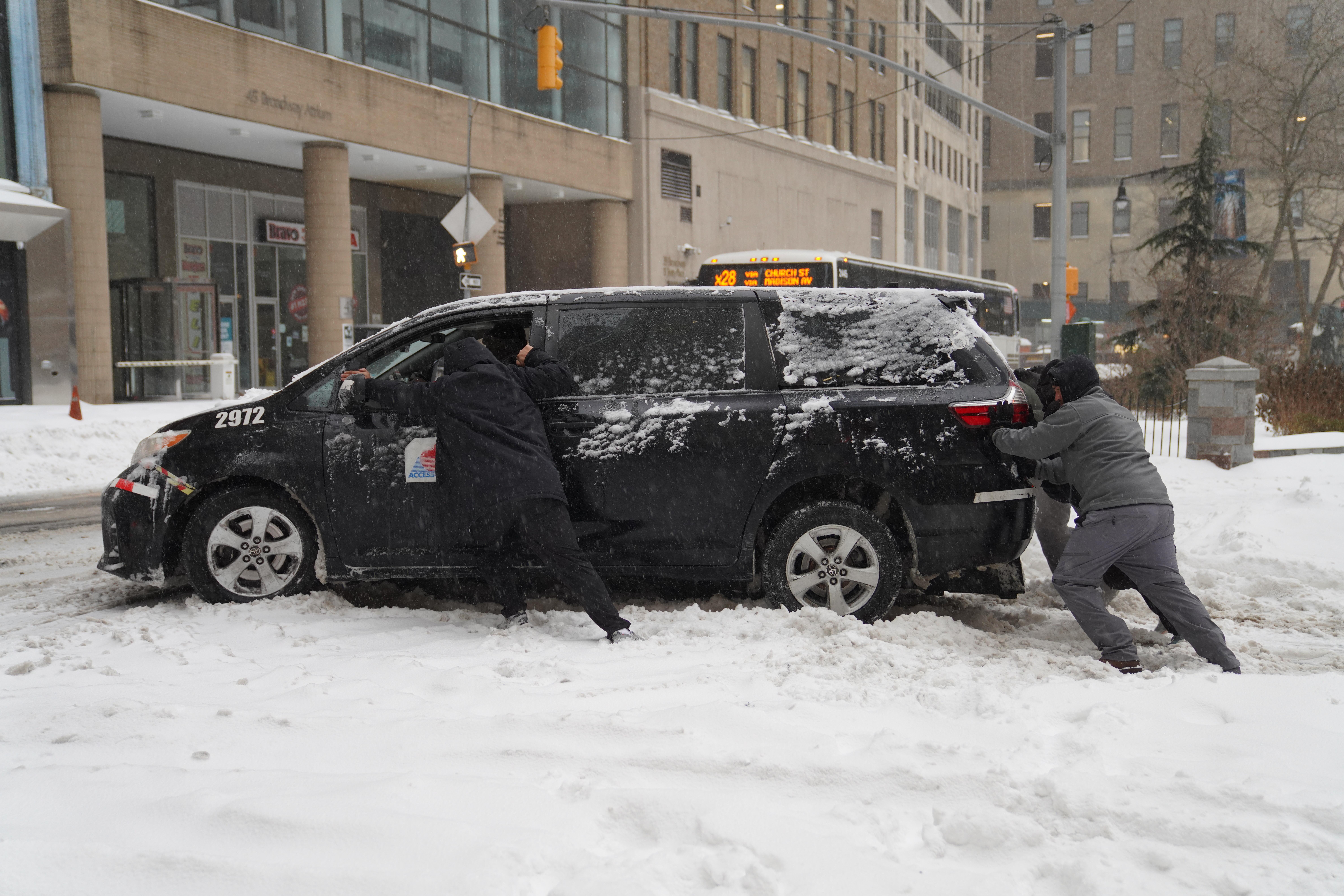 Menschen schieben in New York ein im Schnee festgefahrenes Auto.