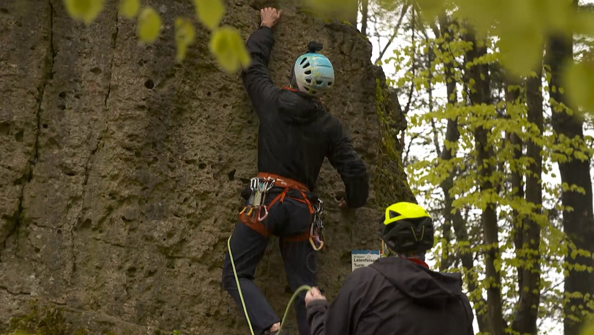 Ein Sportkletterer, gesichert mit einem Seil, klettert auf einen Felsen. 