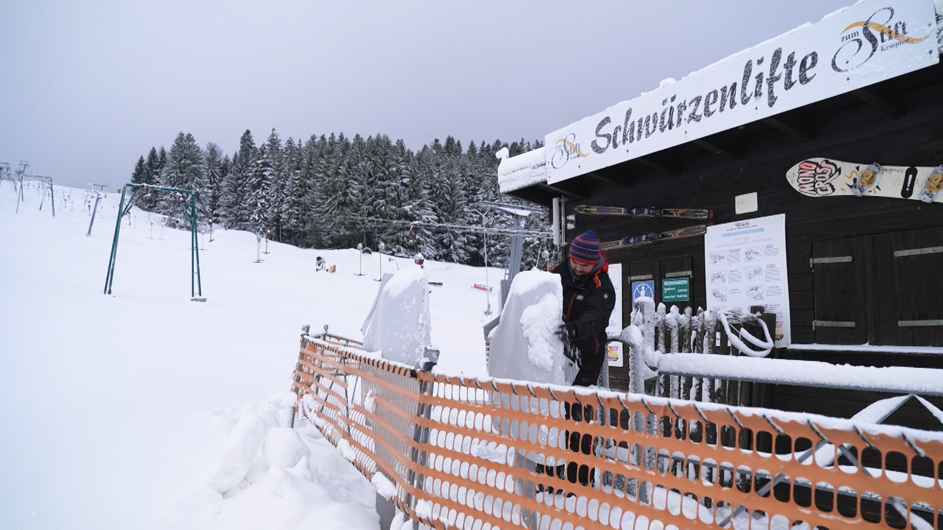 Ein Skilifthäuschen, davor steht ein Mann mit Mütze und klopft das Eis von den Kartenlesern, im Hintergrund der Schlepplift, der noch steht.