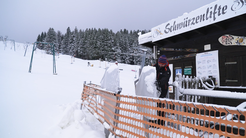 Ein Skilifthäuschen, davor steht ein Mann mit Mütze und klopft das Eis von den Kartenlesern, im Hintergrund der Schlepplift, der noch steht. | Bild: BR / Johannes Hofmann Ein Skilifthäuschen, davor steht ein Mann mit Mütze und klopft das Eis von den Kartenlesern, im Hintergrund der Schlepplift, der noch steht.