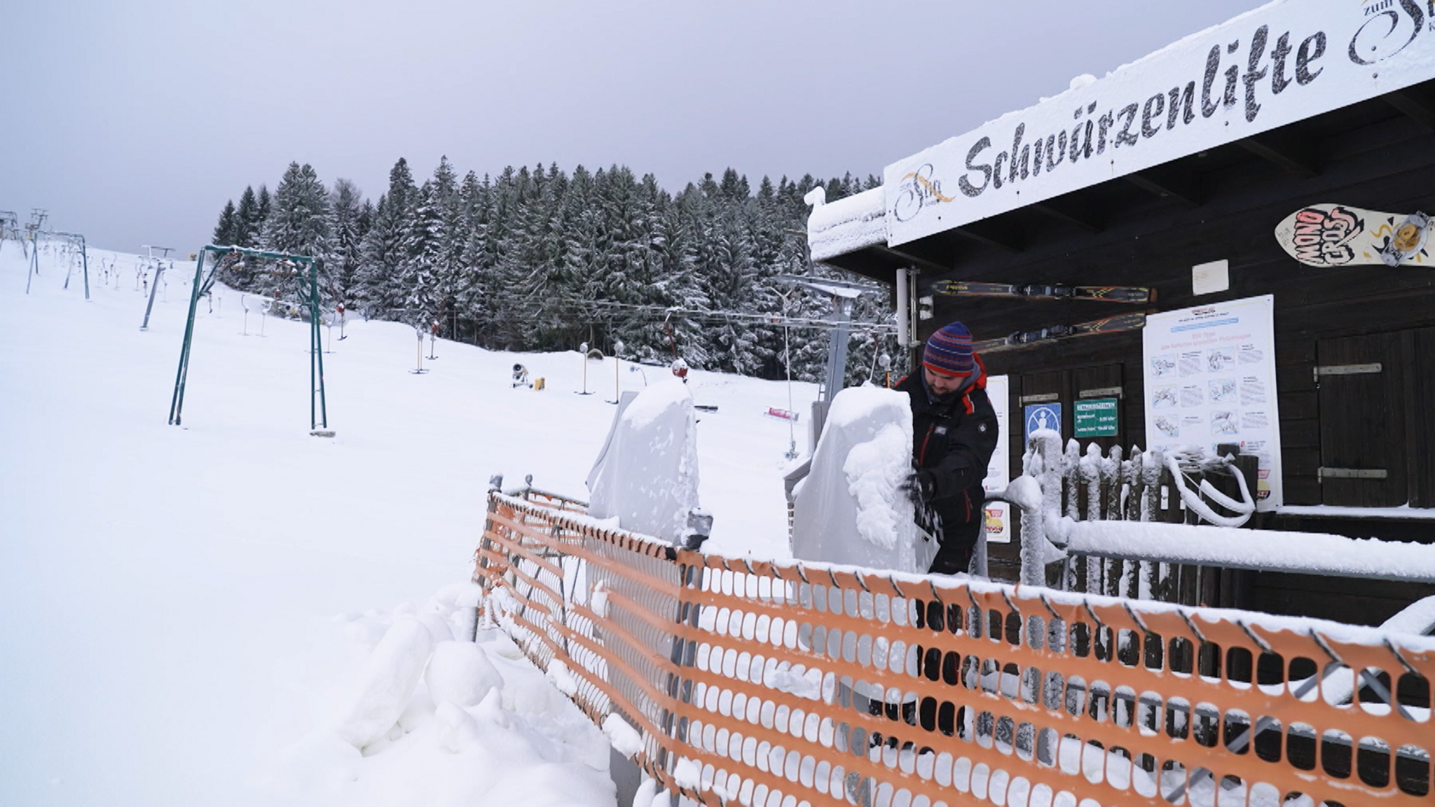 Ein Skilifthäuschen, davor steht ein Mann mit Mütze und klopft das Eis von den Kartenlesern, im Hintergrund der Schlepplift, der noch steht.