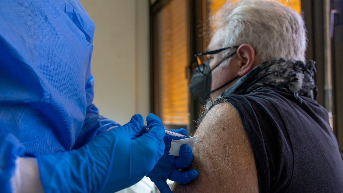 Ein medizinischer Mitarbeiter in blauer Schutzmontur impft einen Mann in den Oberarm.