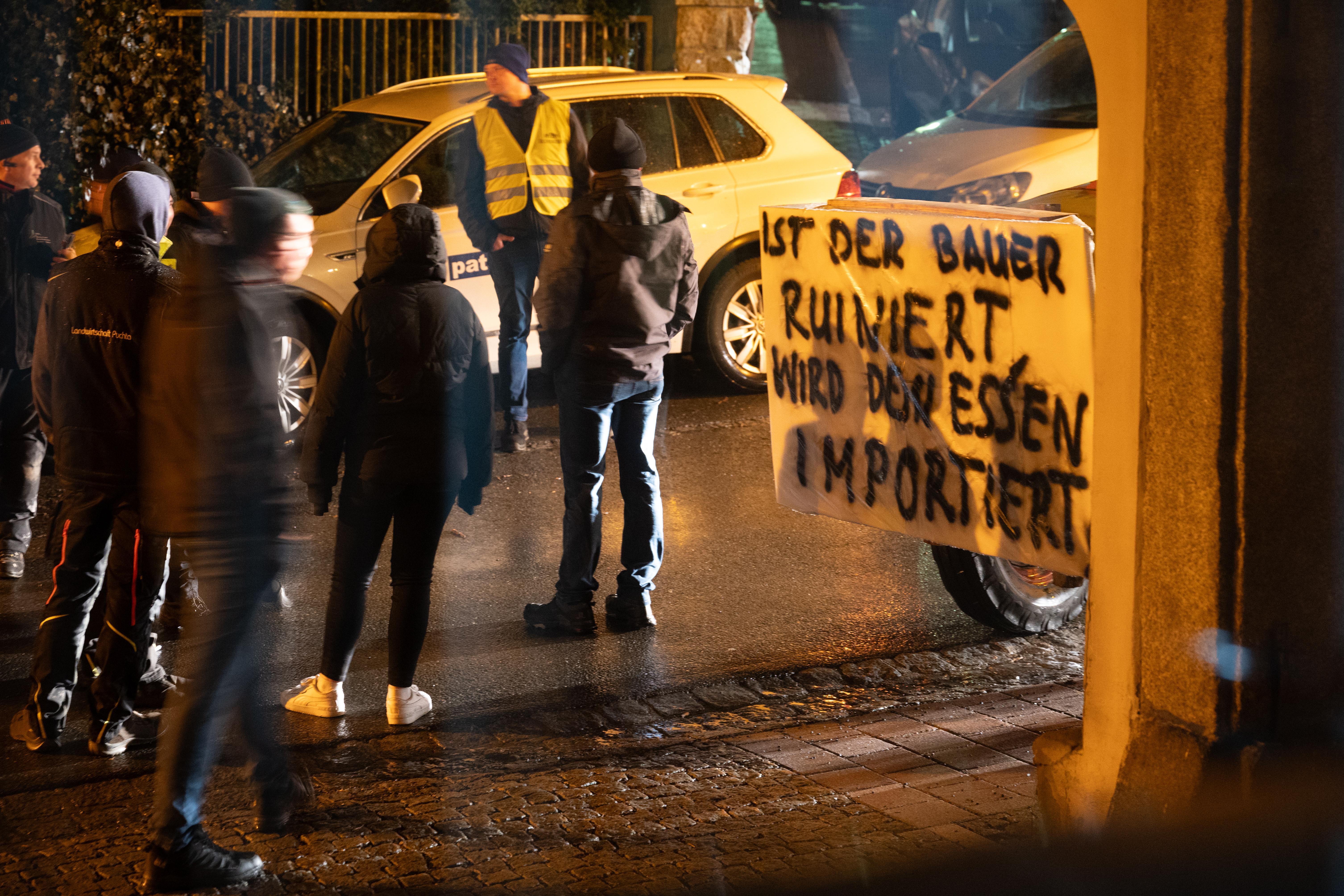 Mehrere Personen stehen auf einer Straße, im Vordergrund steht ein Traktor mit einem Plakat. Es trägt die Aufschrift "Ist der Bauer ruiniert, wird das Essen importiert." 