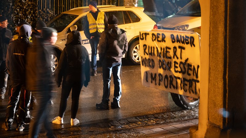 Mehrere Personen stehen auf einer Straße, im Vordergrund steht ein Traktor mit einem Plakat. Es trägt die Aufschrift "Ist der Bauer ruiniert, wird das Essen importiert." 