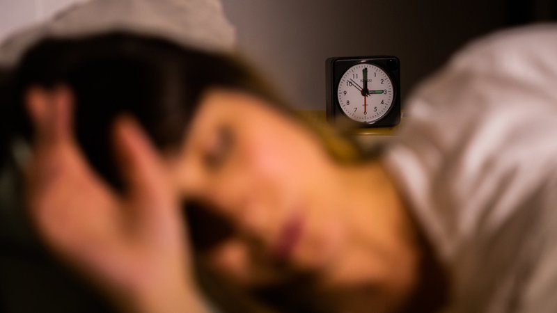 Eine Frau schläft neben einem Wecker, der auf drei Uhr zeigt (gestellte Szene)