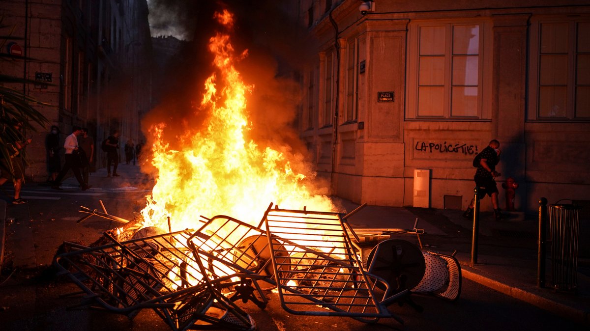 Ein Mensch geht neben einer brennenden Barrikade an einer Mauer vorbei, auf der "Die Polizei tötet" steht.