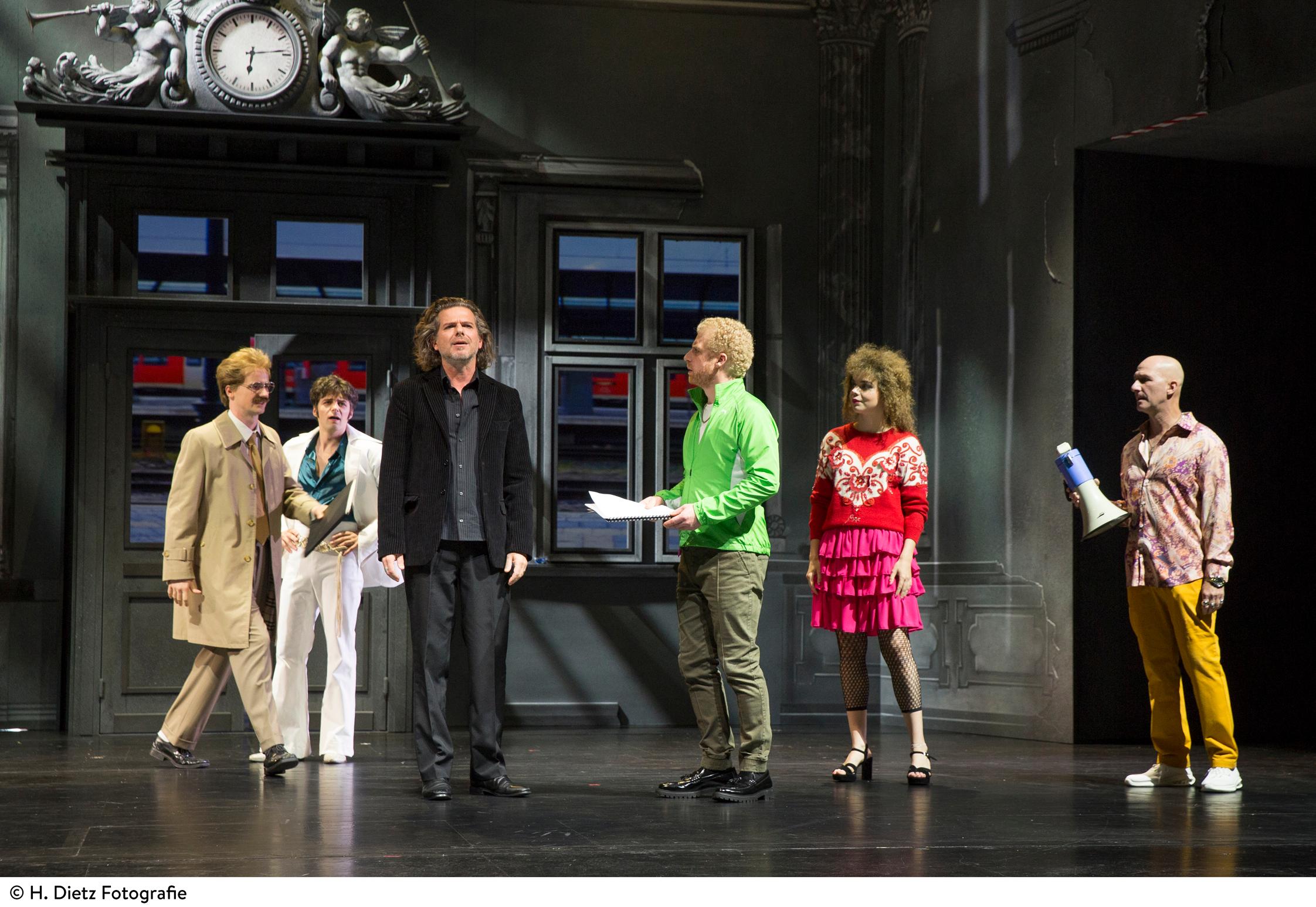 Sechs Schauspieler stehen auf der Bühne im Hofer Theater. Die Kulisse zeigt den Hofer Hauptbahnhof. 