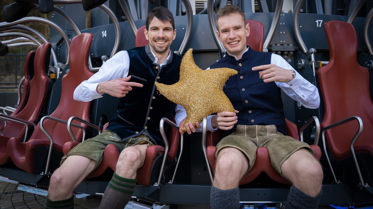 Zwei Männer sitzen in einem Fahrgeschäft im Bayernpark und halten einen Stern, Symbol für die Sternstunden, einer Benefizaktion mit dem Bayerischen Rundfunk.