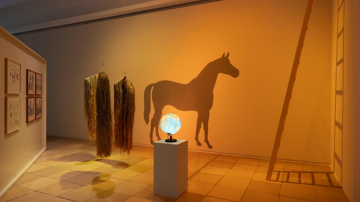 Ein in gelbes Licht getauchter Raum, in dem ein Globus auf einem Sockel steht, dahinter ein Pferd an der Wand projiziert