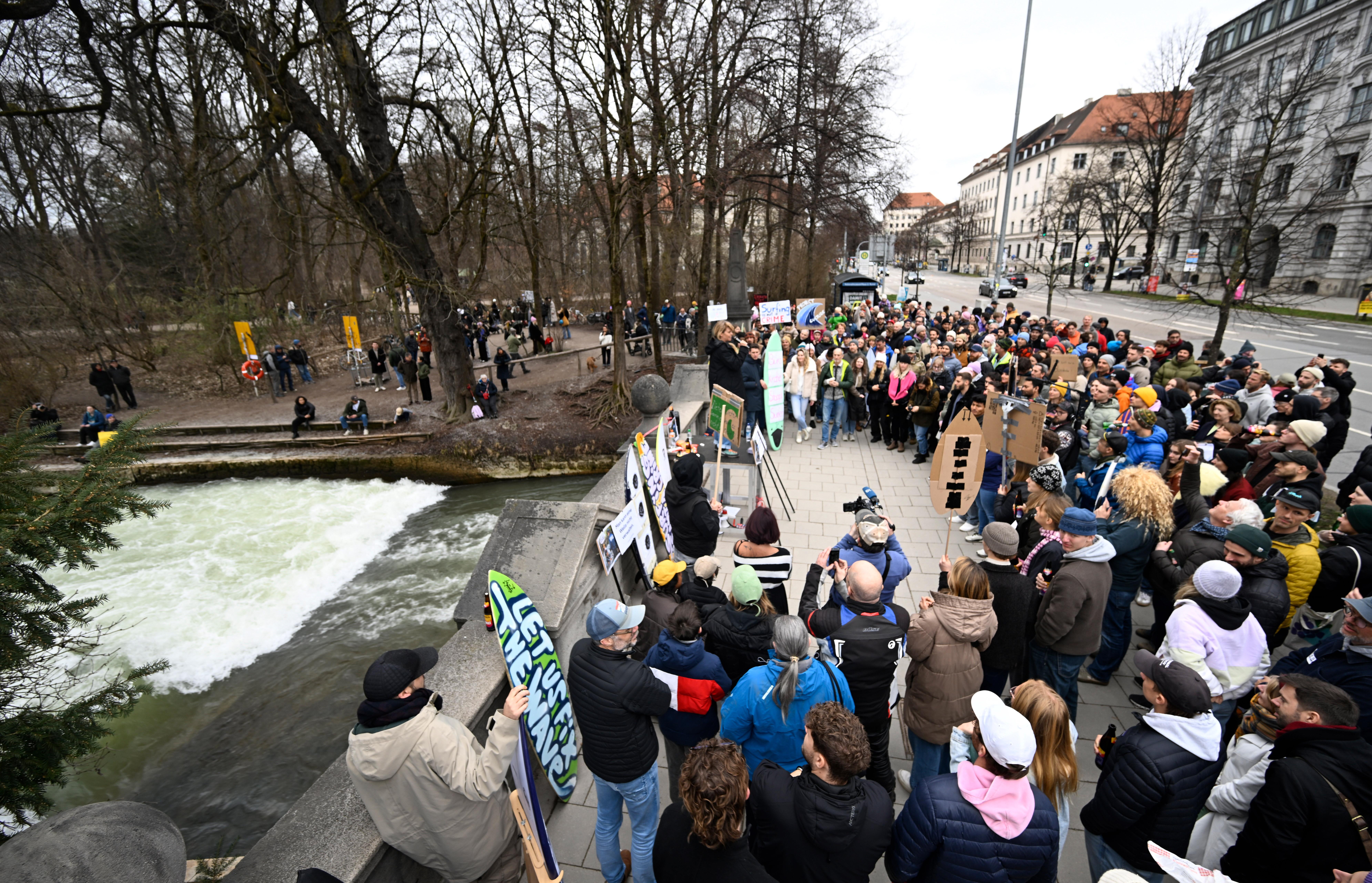 An der Eisbachwelle stehen zahlreiche Protestierende, teilweise mit Pappschildern