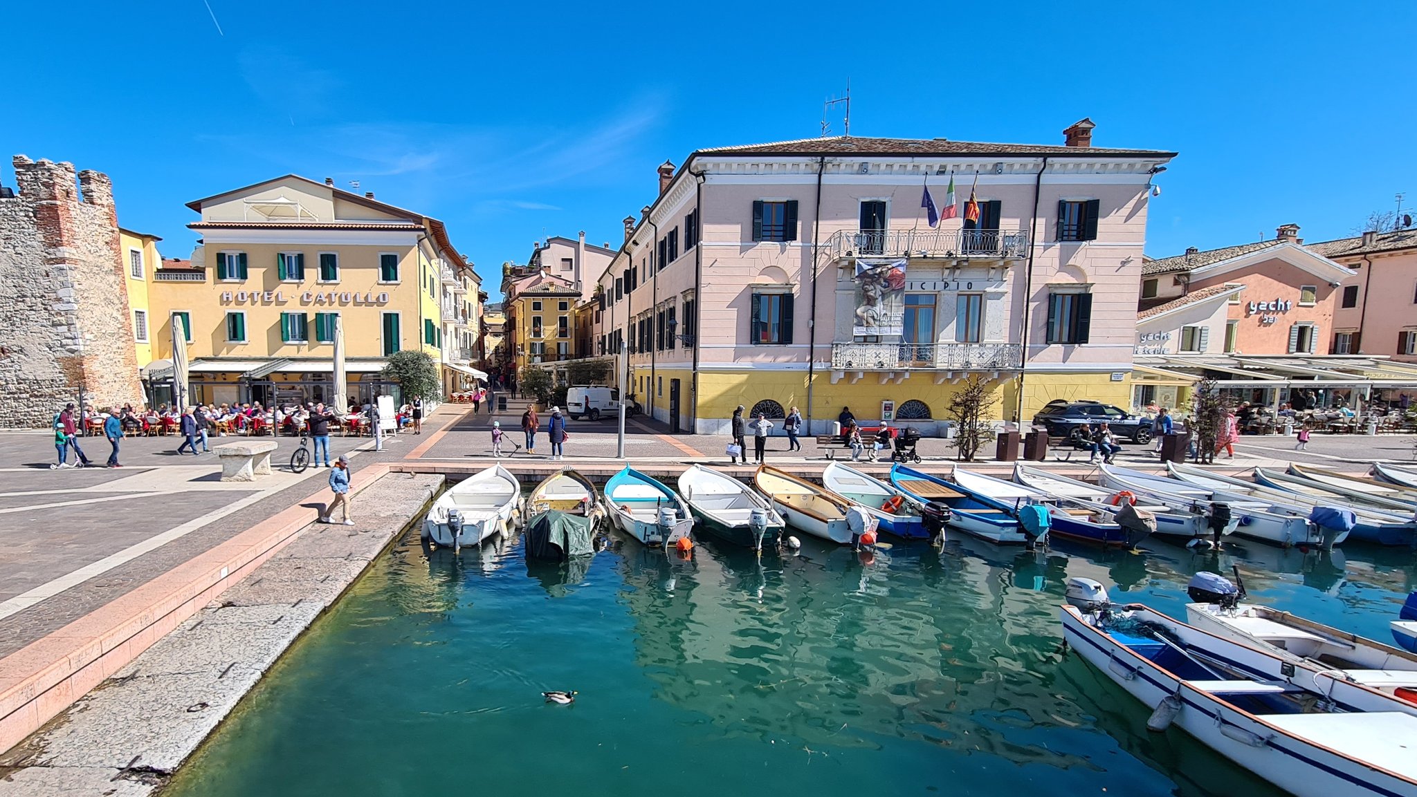 Das Hafenbecken von Bardolino am Gardasee. Fischerboote reihen sich im Wasser aneinander. Menschen sind auf dem Platz unterwegs, im Hintergrund Gebäude vor blauem Himmel. Der Gardasee hat derzeit so viel Wasser wie lange nicht.