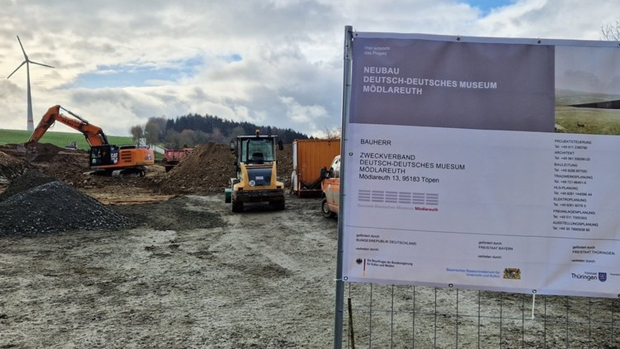 Auf einer Baustelle arbeiten Bagger und Raupen, eine Plakatwand mit der Aufschrift "Neubau Deutsch-Deutsches Museum Mödlareuth" ist aufgestellt. 