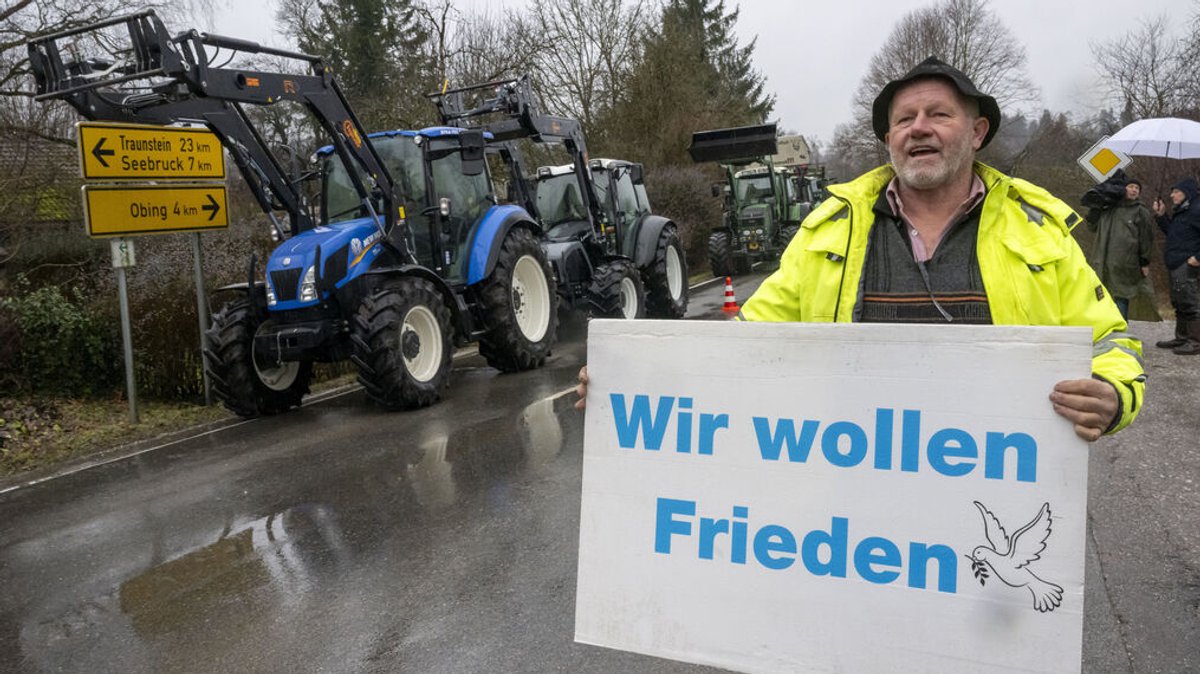 Landwirte haben ihre Traktoren auf einer Zufahrtstraße zum Kloster Seeon abgestellt. Ein Mann hält ein Schild mit der Aufschrift "Wir wollen Frieden".