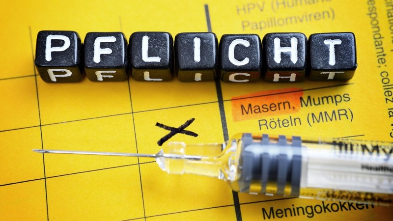 Impfspritze auf einem gelben Impfausweis, bei dem die Masern markiert sind. Würfel mit Buchstaben "Pflicht" liegen darauf.