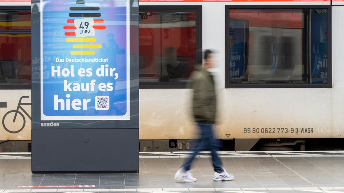 Ein junger Mann geht an einer Bahn vorbei. Daneben steht ein digitales Werbeplakat zum Deutschlandticket.