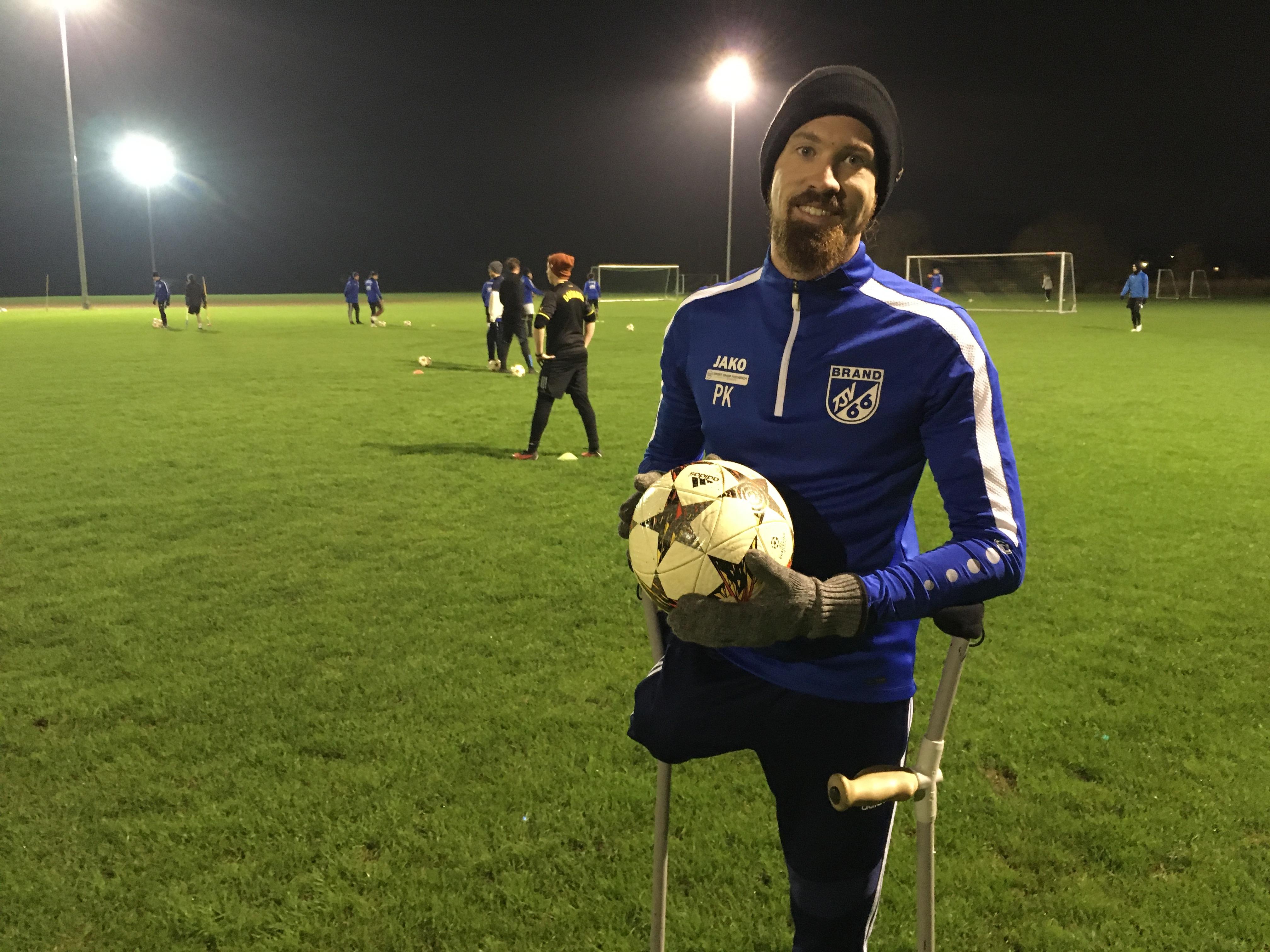 Pierre Kaiser steht an Krücken mit einem Fußball in der Hand auf einem Fußballplatz.