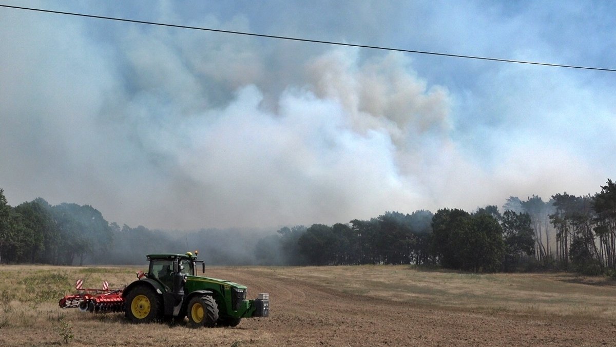 Ein Traktor steht auf einem Feld vor einem Waldstück, aus dem Rauchwolken aufsteigen.