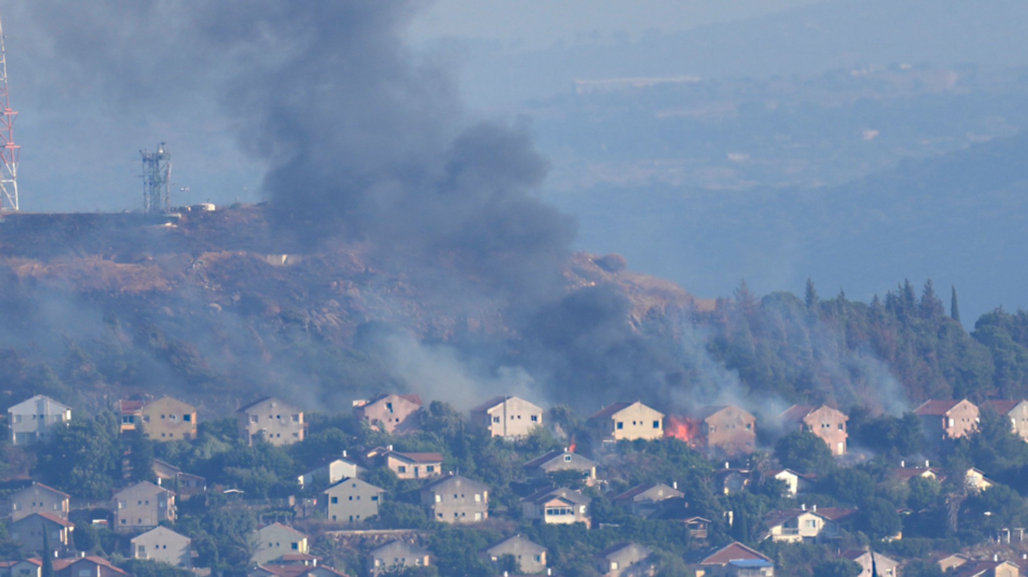 22.06.2024, Libanon, Marjayoun: Feuer und schwarzer Rauch steigen zwischen den Häusern der nördlichen israelischen Grenzstadt Metula auf, die von der Hisbollah beschossen wurde, fotografiert aus Marjayoun im Südlibanon.