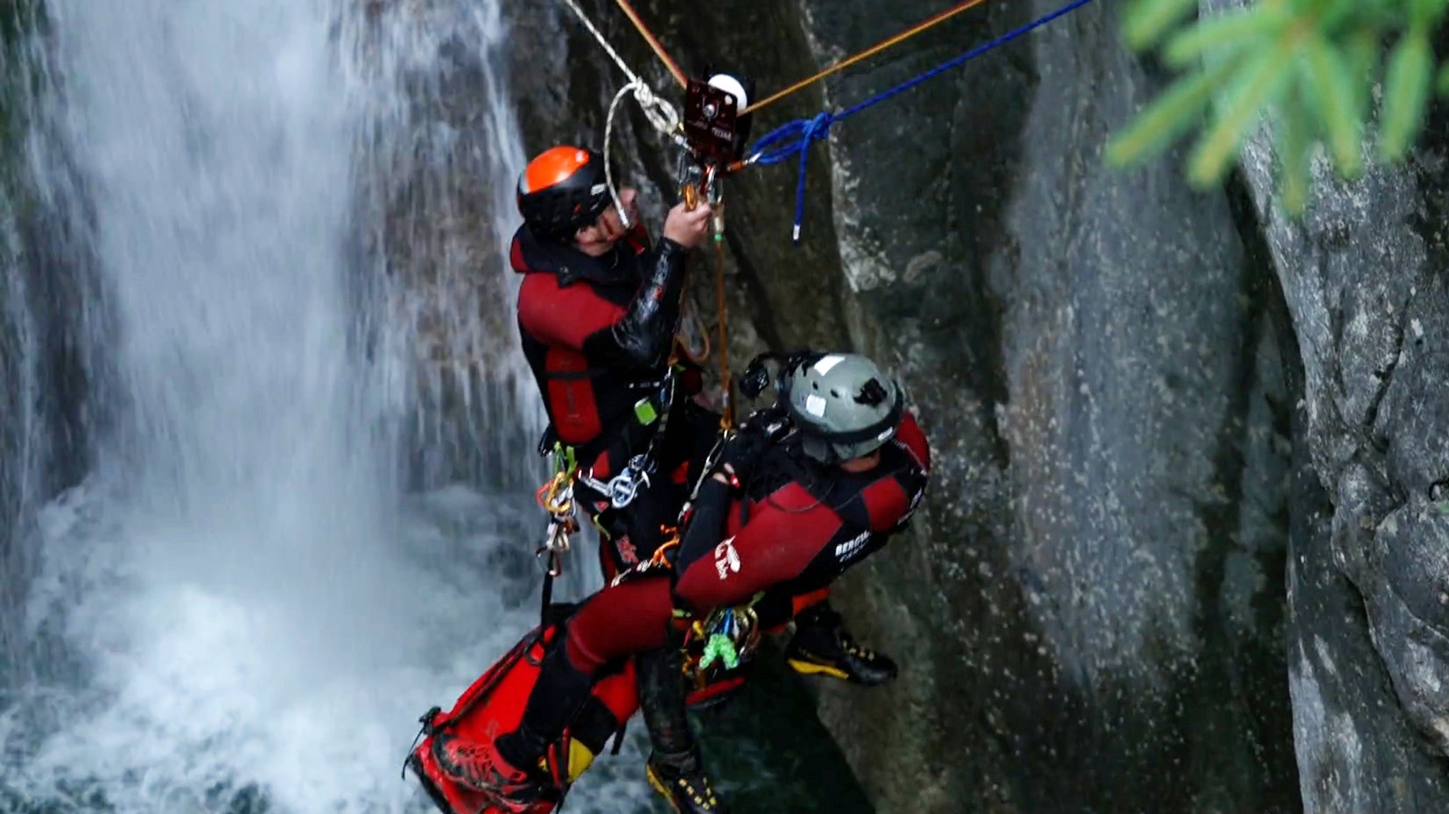 Zwei Bergretter in Neoprenanzug hängen an Seilen vor einem Wasserfall