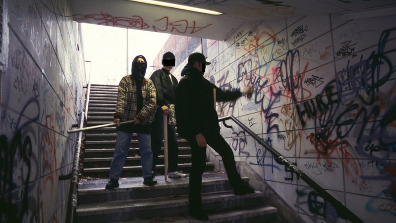 Mitglieder einer Jugendbande mit einer Eisenstange und einer Holzlatte stehen in einem Tunnel, ein Jugendlicher besprüht die Wand mit Graffitis.