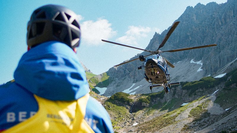 Symboldbild_ Ein Bergwacht-Retter im Vordergrund, dahinter ein schwebender Hubschrauber in Bergkulisse | Bild: BR/Luis Trautmann Symboldbild_ Ein Bergwacht-Retter im Vordergrund, dahinter ein schwebender Hubschrauber in Bergkulisse
