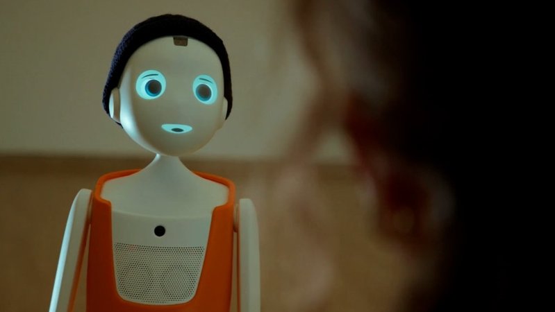 Ein kleiner Sozialroboter mit orangenem Körper schaut nach vorne. Im Vordergrund sitzt eine Frau | Bild: BR Ein kleiner Sozialroboter mit orangenem Körper schaut nach vorne. Im Vordergrund sitzt eine Frau