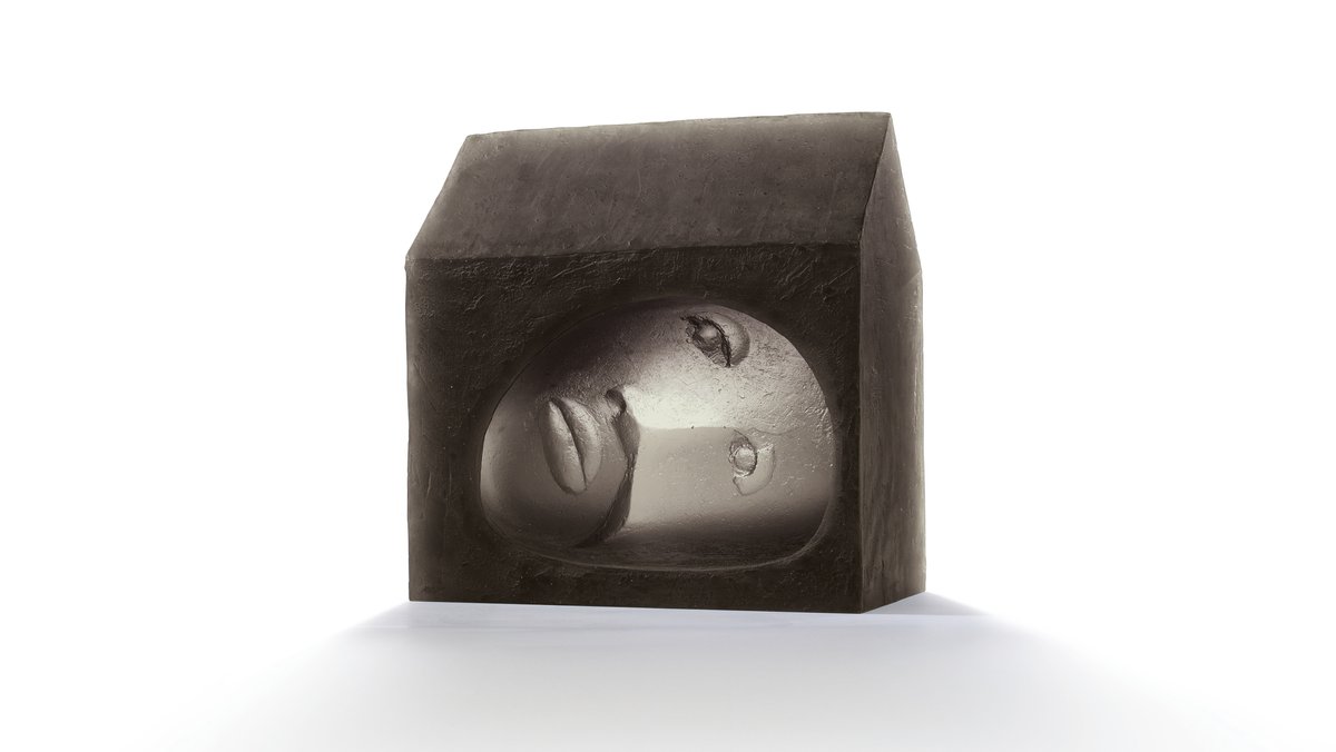 Stilisierter Kopf in Grau in einem hausförmigen, massiven Bock aus schwarzem Glas