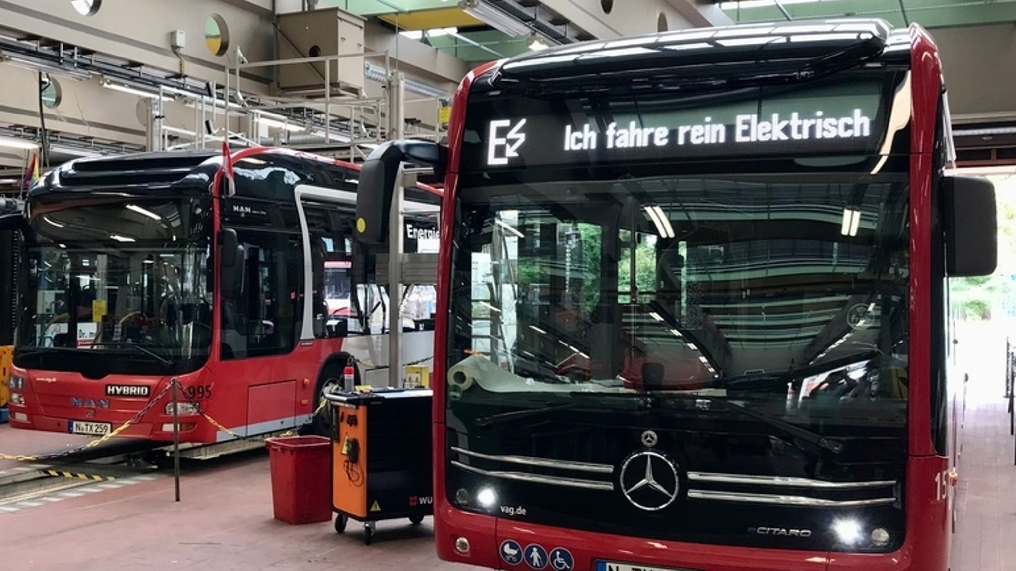 Zwei rote Busse stehen in einer Halle mit der Hinweisanzeige "Ich fahre elektrisch". | Bild: BR/ Michael Reiner Zwei rote Busse stehen in einer Halle mit der Hinweisanzeige "Ich fahre elektrisch".