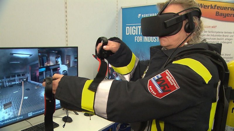 Feuerwehrfrau testet eine VR-Brille vor einem Bildschirm.