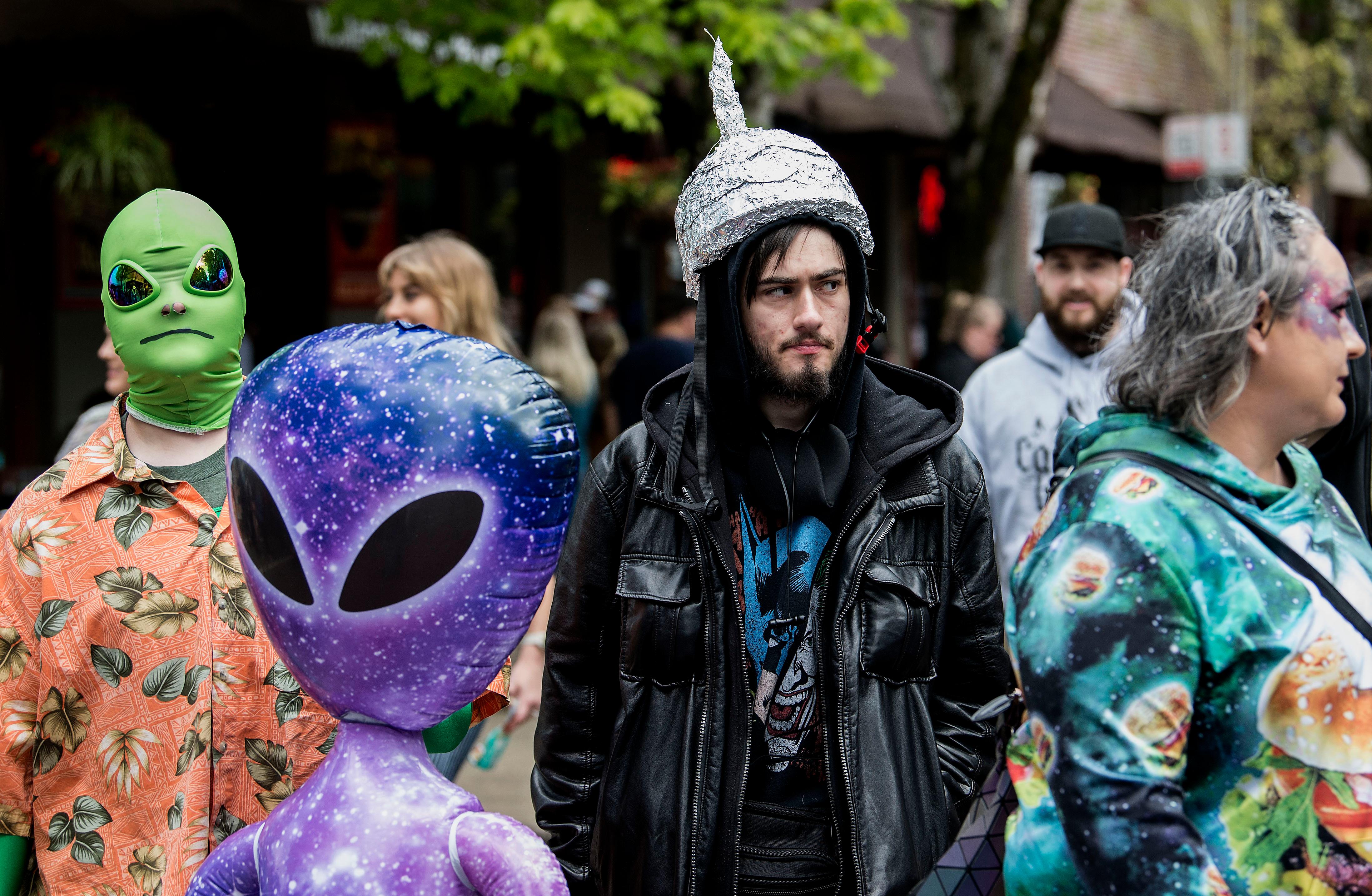 Ein Mann mit einem Hut aus Aluminium-Folie steht vor einem aufblasbaren Alien, links daneben ein Mann mit einer grüner Außerirdischen-Maske auf dem Kopf.