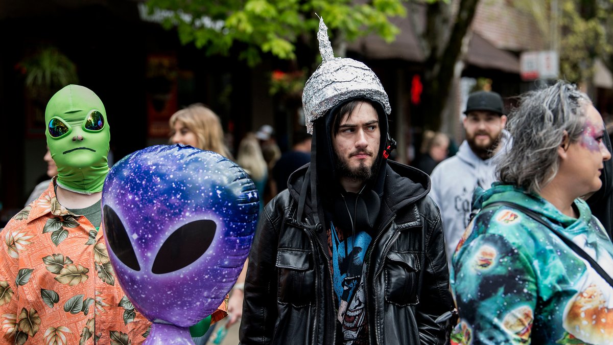 Ein Mann mit einem Hut aus Aluminium-Folie steht vor einem aufblasbaren Alien, links daneben ein Mann mit einer grüner Außerirdischen-Maske auf dem Kopf.