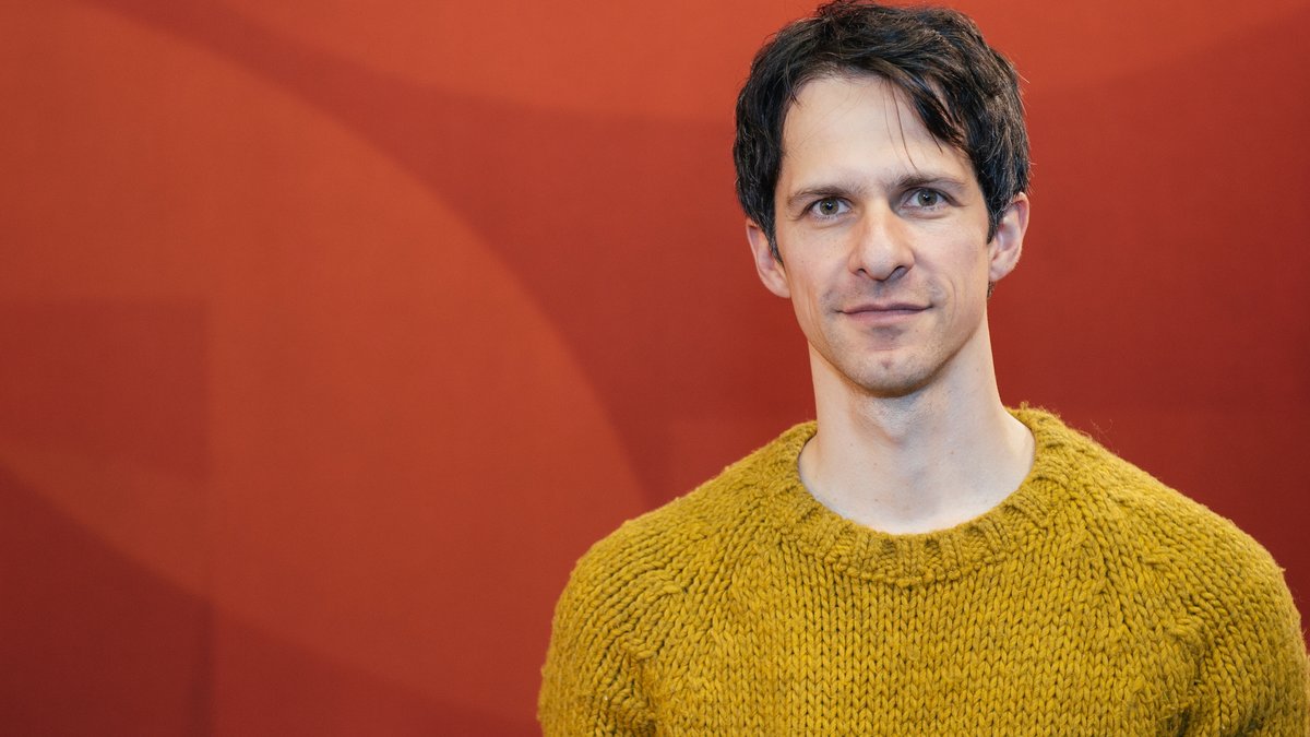 Autor Thomas von Steinaecker steht in gelbem Pullover vor einer roten Wand und blickt freundlich in die Kamera
