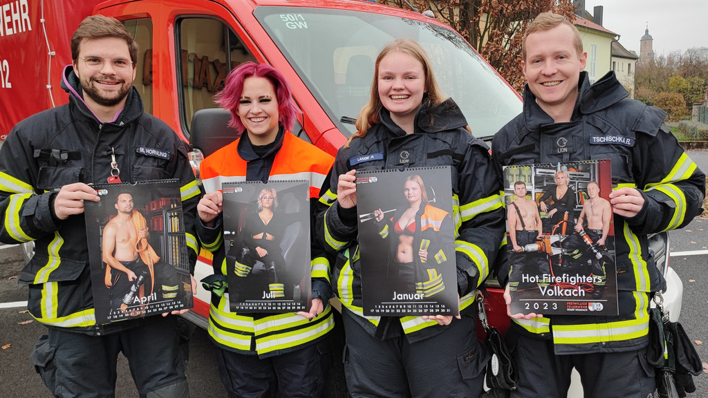 Vier Mitglieder der Freiwilligen Feuerwehr Volkach, zwei Frauen und zwei Männer, stehen in Feuerwehr-Montur vor einem Feuerwehrauto. Jeder hält einen Kalender in den Händen. Aufgeschlagen ist das jeweilige Kalenderblatt, auf dem sie selbst zu sehen sind - obenrum nicht oder nur wenig bekleidet.