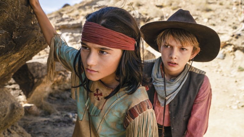 Filmbild aus "Der junge Häuptling Winnetou": Ein indigener Junge und ein weißer Junge mit Cowboyhut blicken suchend um einen Felsen