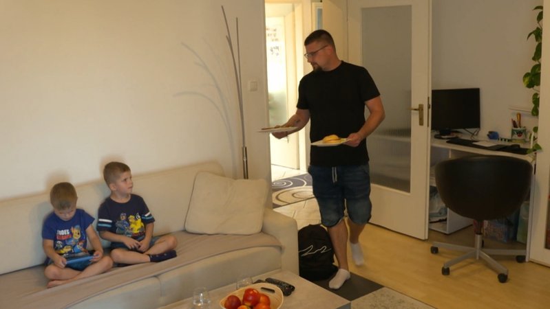 Ein Mann kommt mit zwei Tellern in ein Wohnzimmern, auf dem Sofa sitzen seine Söhne.