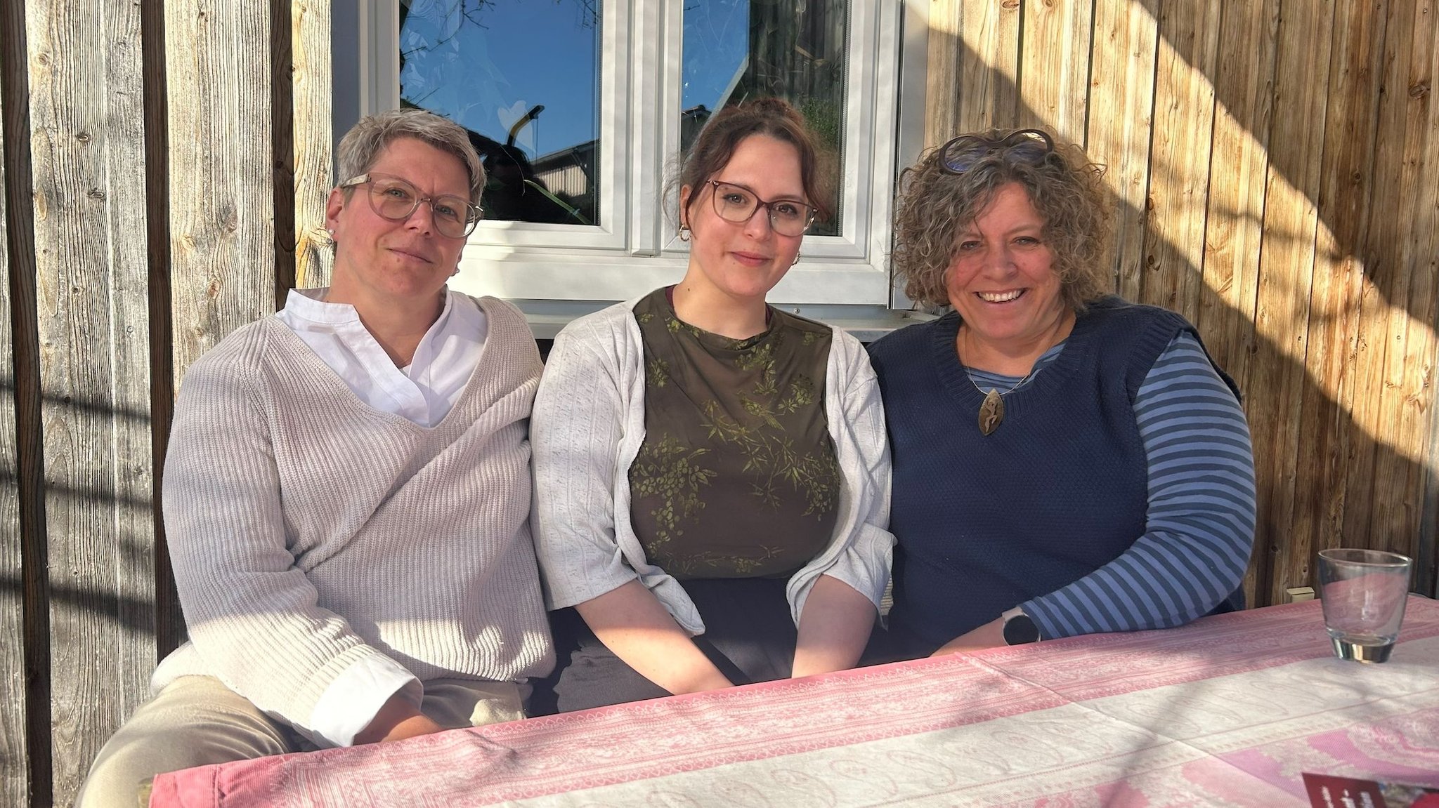 Drei Frauen sitzen auf einer Bank vor einem Holzhaus in einem Garten 