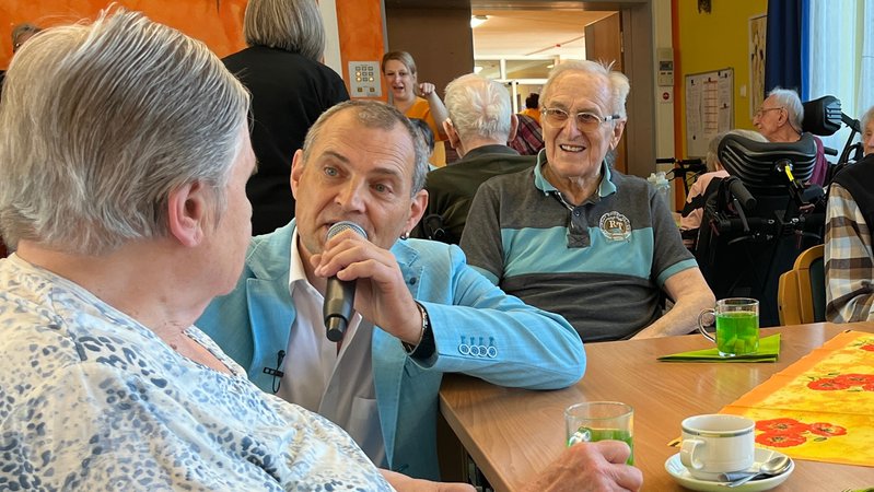 Pflegedienstleiter Norbert Köhler kniet neben einer Seniorin am Tisch und singt.