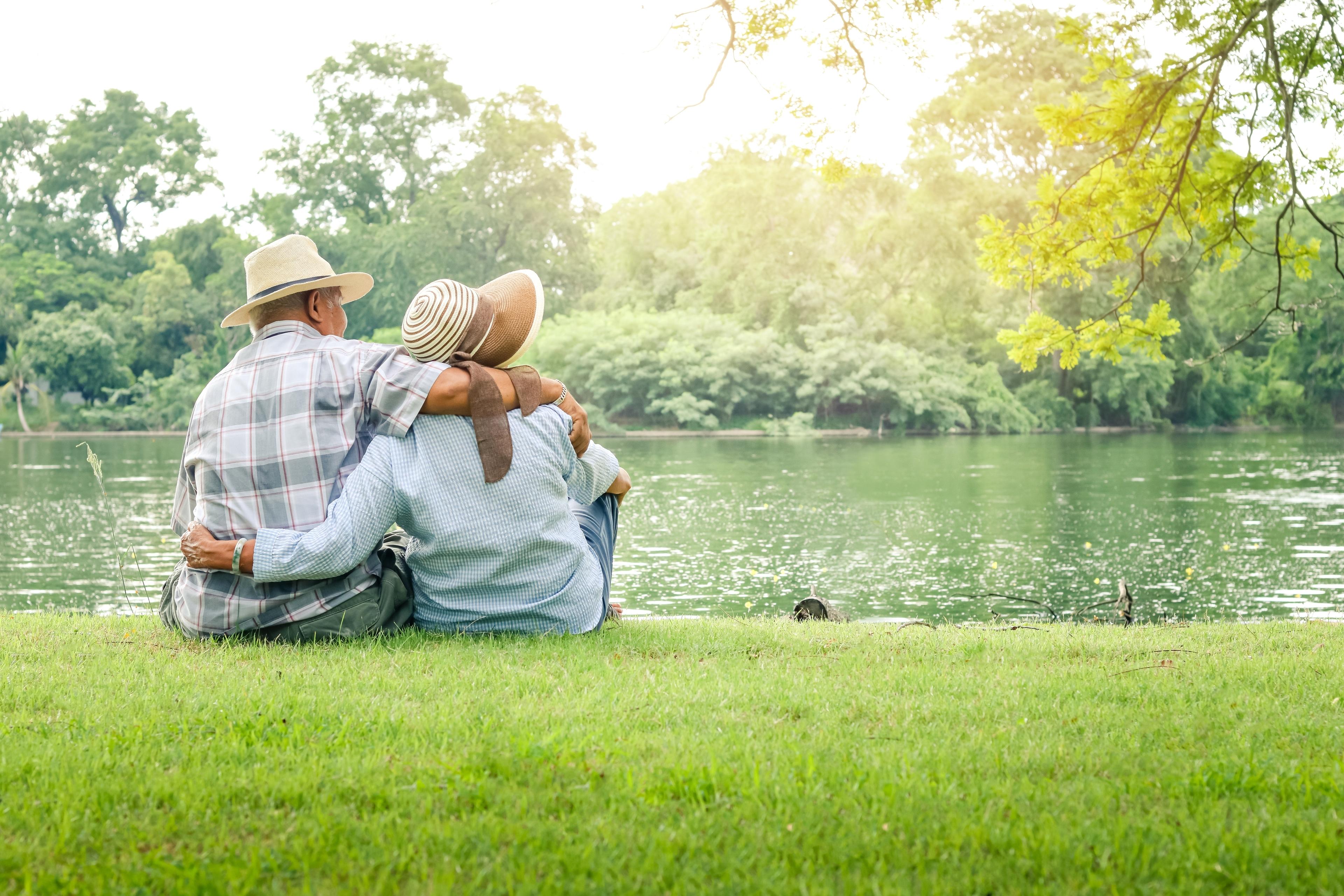 Ein älteres Paar sitzt eng umschlungen auf einer Wiese am Ufer eines ruhigen Sees. Eine Studie zeigt, dass gemeinsam erlebte Freude das Stresshormon Cortisol senkt und damit gesundheitsfördernd ist.