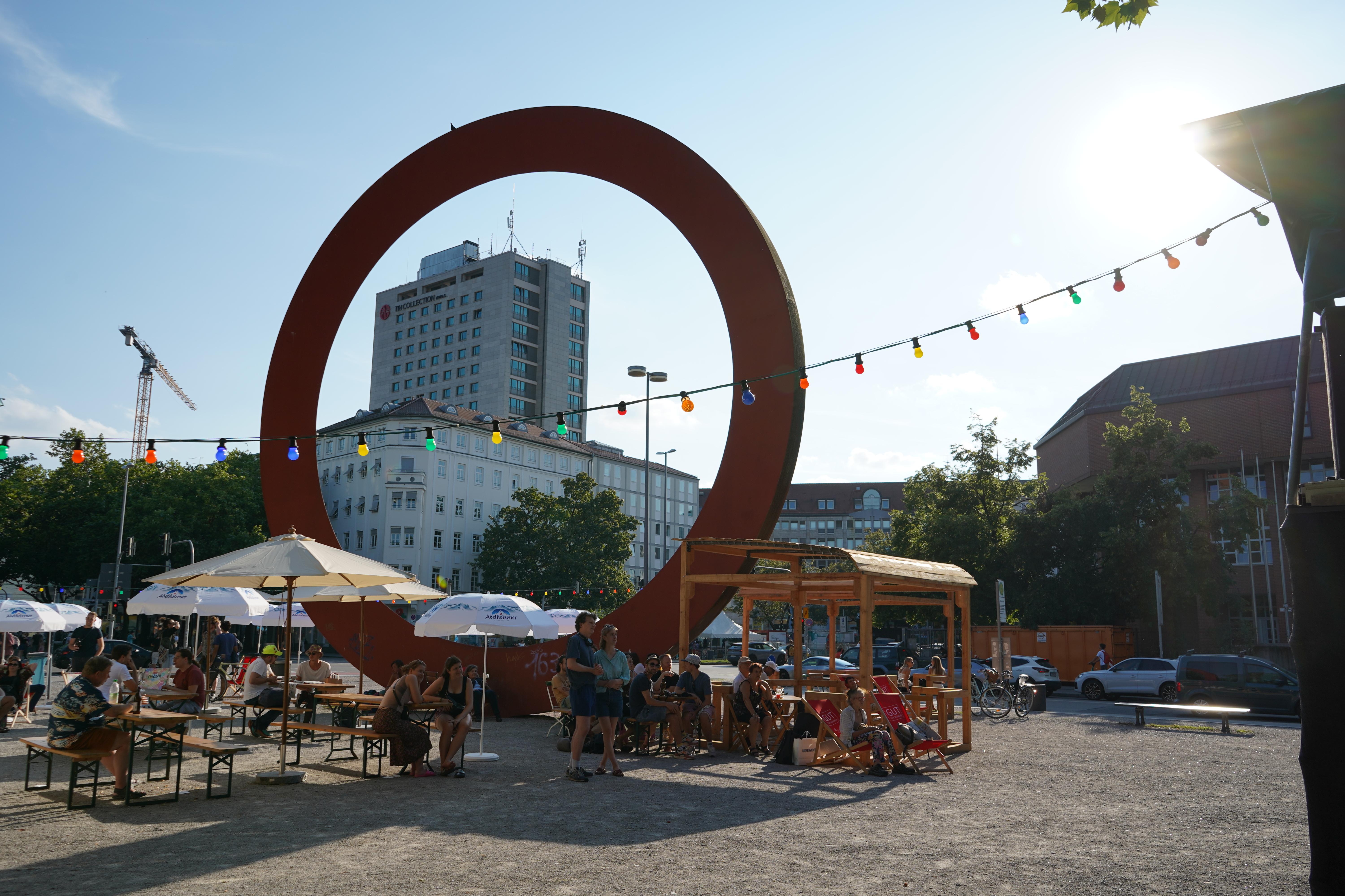 Bierbänke und Sonnenschirme auf einem gepflasterten Platz. Im Hintergrund ein 12 Meter großer Ring, Kunstwerk, durch den man Häuser sieht. als Kunstwerk rund