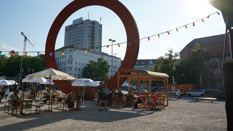 Bierbänke und Sonnenschirme auf einem gepflasterten Platz. Im Hintergrund ein 12 Meter großer Ring, Kunstwerk, durch den man Häuser sieht. als Kunstwerk rund | Bild: "Die Null" I Corinna Böck Bierbänke und Sonnenschirme auf einem gepflasterten Platz. Im Hintergrund ein 12 Meter großer Ring, Kunstwerk, durch den man Häuser sieht. als Kunstwerk rund