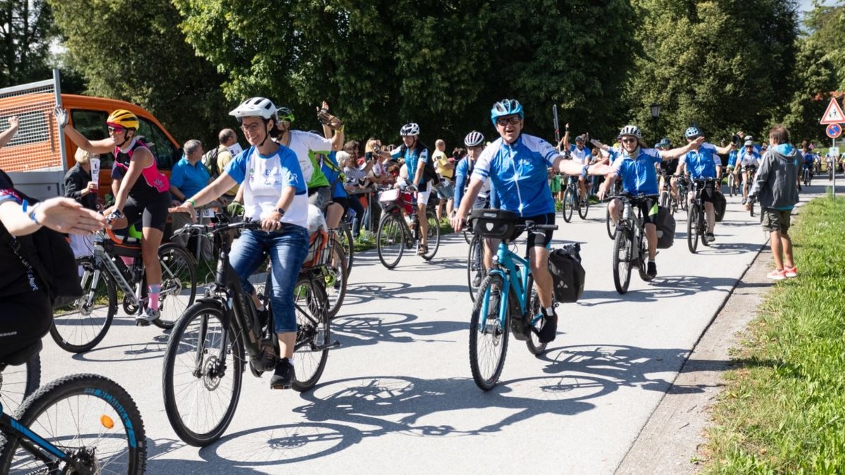 Hobbyradlerinnen und – radler fahren bei der BR-Radltour 2023 bei sonnigem Wetter eine Straße entlang und winken fröhlich in die Kamera. Dabei tragen einige blau-weiße Trikots.